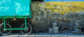 A large turquoise metal cart with two bicycle wheels is parked next to an old, weathered wall. The wall features a faded mural of a hand reaching out, painted in black on a yellow background. Several stacked stones rest on the ground against the wall.
