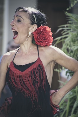 A flamenco dancer passionately performing, with her mouth open as if singing or expressing strong emotion. She is adorned with a large red rose in her hair and is wearing a black and red dress with a fringe. Her hair is slicked back, and she has pearl earrings. The background features some greenery.