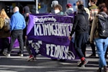 A group of people is participating in a protest or march, holding a purple banner with white text that promotes feminism and climate justice. Some individuals are wearing masks, and they are dressed in casual attire. The environment is outdoor, possibly on a city street.