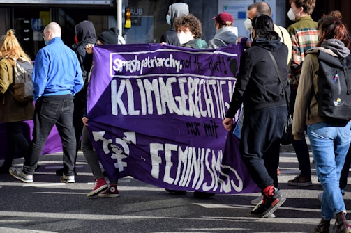 A group of people is participating in a protest or march, holding a purple banner with white text that promotes feminism and climate justice. Some individuals are wearing masks, and they are dressed in casual attire. The environment is outdoor, possibly on a city street.