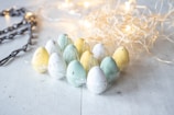 A dozen pastel-colored speckled eggs are arranged in a plastic egg carton. The colors include shades of yellow, green, and white. Decorative string lights and straw-like material are arranged in the background, creating a soft and festive ambiance.