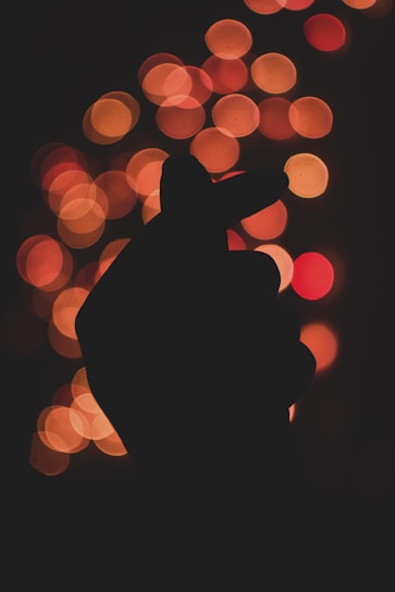 A silhouetted hand forms a heart shape with its fingers against a background of warm, out-of-focus bokeh lights in shades of red, orange, and yellow.