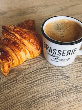 A croissant with a golden-brown crust sits next to a mug of coffee on a wooden table. The mug is white with black lettering that reads 'Brasserie Cooking Company.' The table has a textured wood grain finish, adding a rustic feel to the setting.