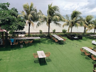 A beachfront outdoor dining area with several long wooden tables surrounded by wicker chairs on a grassy surface. Palm trees line the edge of the area, overlooking a body of water. String lights are strung across the space, creating a cozy atmosphere. A person wearing a blue shirt and mask is walking under the tree canopy on the left side of the image.