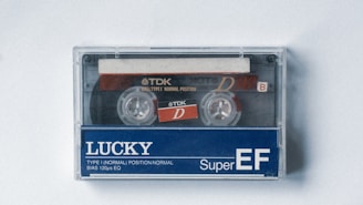 A vintage audio cassette tape displayed inside a transparent plastic case, featuring a black tape reel and a label with the brand TDK. The bottom half of the cassette is prominently labeled 'LUCKY Super EF' with additional text indicating type and position specifications.