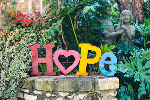 Colorful letters spelling the word 'hope' are arranged on a stone surface amidst lush green foliage. The 'O' is creatively designed as a heart. Behind the letters, two stone statues of children are partly visible, surrounded by dense vegetation.