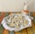 A plush bunny toy with an orange bow sits next to a plate filled with bunny-shaped cookies, decorated with white icing. The cookies are arranged on a white cloth within the plate, which rests on a colorful woven tablecloth.