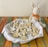 A plush bunny toy with an orange bow sits next to a plate filled with bunny-shaped cookies, decorated with white icing. The cookies are arranged on a white cloth within the plate, which rests on a colorful woven tablecloth.