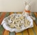 A plush bunny toy with an orange bow sits next to a plate filled with bunny-shaped cookies, decorated with white icing. The cookies are arranged on a white cloth within the plate, which rests on a colorful woven tablecloth.