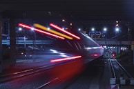 A high-speed train moves swiftly under a bridge at night, with bright red and white lights creating a dramatic motion blur effect. The scene is illuminated by overhead street lights and the city skyline, contributing to a dynamic urban atmosphere.