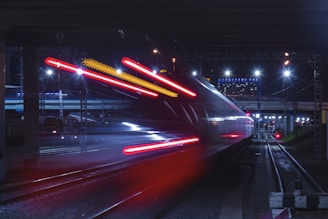 A high-speed train moves swiftly under a bridge at night, with bright red and white lights creating a dramatic motion blur effect. The scene is illuminated by overhead street lights and the city skyline, contributing to a dynamic urban atmosphere.