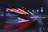 A high-speed train moves swiftly under a bridge at night, with bright red and white lights creating a dramatic motion blur effect. The scene is illuminated by overhead street lights and the city skyline, contributing to a dynamic urban atmosphere.