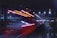 A high-speed train moves swiftly under a bridge at night, with bright red and white lights creating a dramatic motion blur effect. The scene is illuminated by overhead street lights and the city skyline, contributing to a dynamic urban atmosphere.