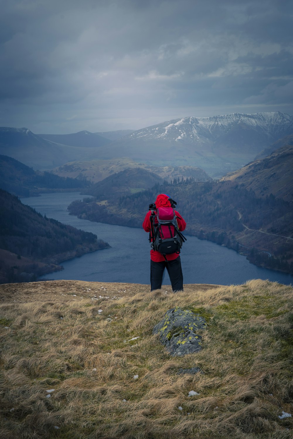 man in red and black jacket standing on brown rock near lake during daytime