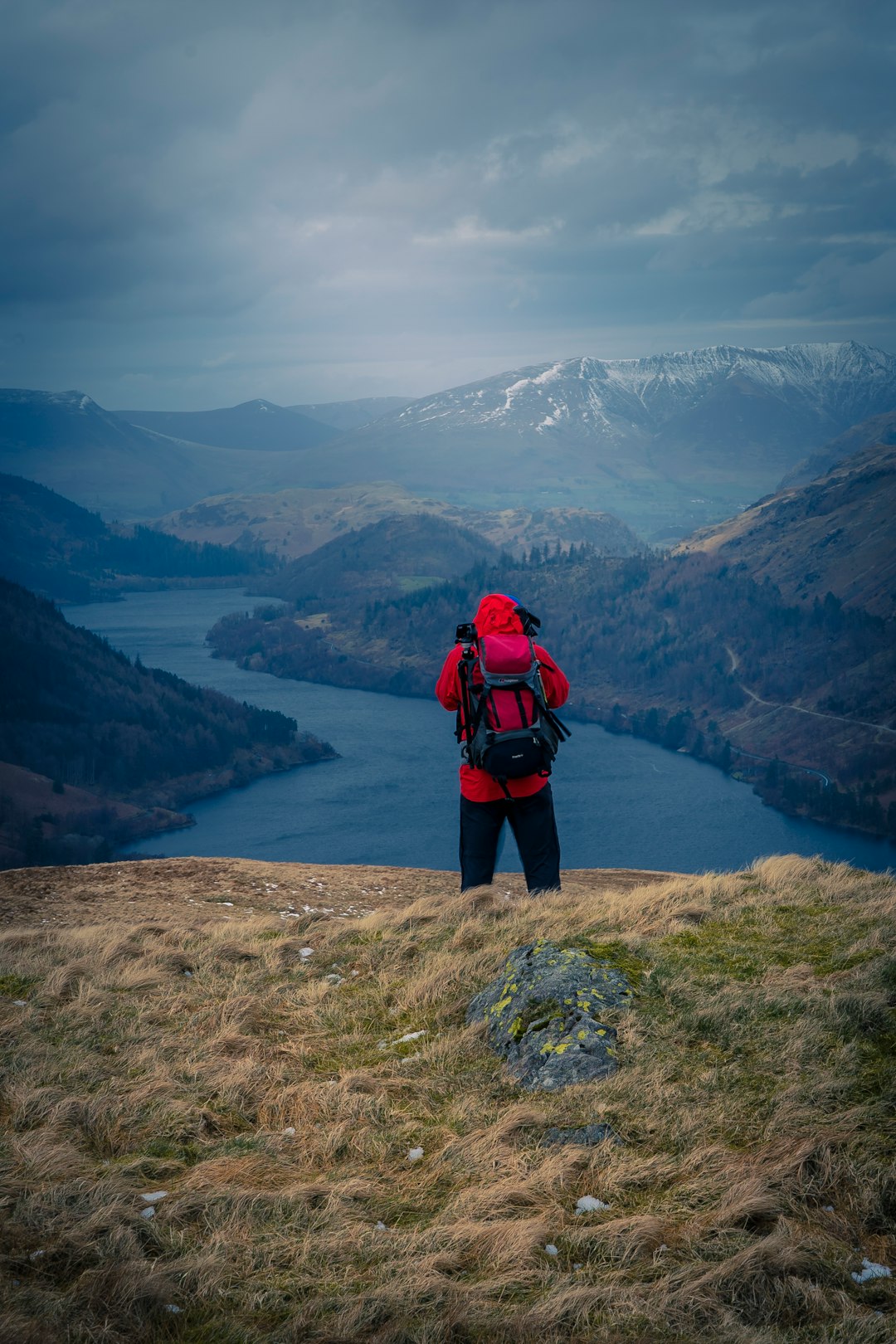 man in red and black jacket standing on brown rock near lake during daytime