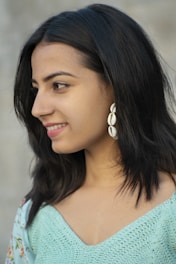 A woman with dark hair and a pleasant smile is captured in profile view. She is wearing a pair of distinctive shell earrings and a light blue, patterned top featuring floral accents.