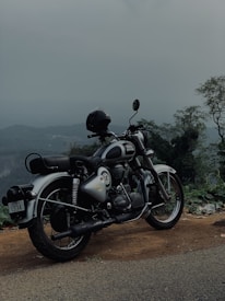 A classic motorcycle parked on a scenic overlook, with a helmet resting on the seat. The backdrop features a misty, overcast sky blending into distant hills and lush greenery, creating a serene and adventurous ambiance.