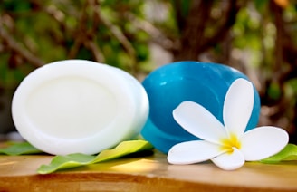 Two oval-shaped soaps, one white and one blue, are placed on green leaves on a wooden surface. A white flower with a yellow center is positioned in front of the blue soap. The background is slightly blurred with green foliage visible.