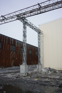 A metal structure with steel beams extends across the sky, connected by sturdy concrete pillars. The background features a weathered building with rust-colored panels and small windows. The foreground is covered with partially melted snow and muddy ground, suggesting a cold and industrial setting.