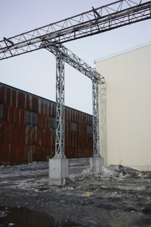 A metal structure with steel beams extends across the sky, connected by sturdy concrete pillars. The background features a weathered building with rust-colored panels and small windows. The foreground is covered with partially melted snow and muddy ground, suggesting a cold and industrial setting.
