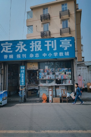 A small newsstand located on the ground floor of a beige building with multiple balconies. The newsstand has a blue sign with white text in a foreign language. The display window shows various newspapers and magazines for sale. A young boy holding a plastic bag is walking past the newsstand on the street. Items such as chairs, a basket, and other objects are placed in front of the store.