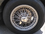 A close-up view of a vintage car wheel, featuring a wire-spoke design and a central hubcap with the Jaguar logo. The tire is labeled with brand and size specifications. The metallic sheen of the spokes contrasts with the dark rubber of the tire.