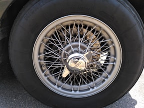 A close-up view of a vintage car wheel, featuring a wire-spoke design and a central hubcap with the Jaguar logo. The tire is labeled with brand and size specifications. The metallic sheen of the spokes contrasts with the dark rubber of the tire.