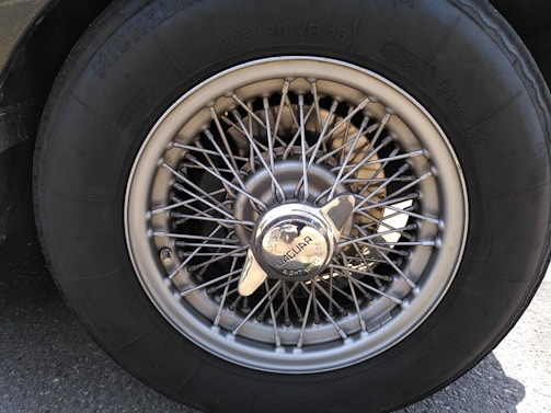 A close-up view of a vintage car wheel, featuring a wire-spoke design and a central hubcap with the Jaguar logo. The tire is labeled with brand and size specifications. The metallic sheen of the spokes contrasts with the dark rubber of the tire.
