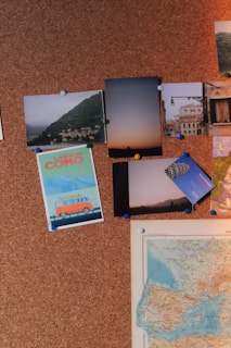 A corkboard with various pinned items including photographs, postcards, and a map. The photos depict scenic landscapes, city streets, and an illustration of a van with the text 'Lago di Como'. The map shows a section of what appears to be Europe. It is organized with colored push pins holding everything in place.