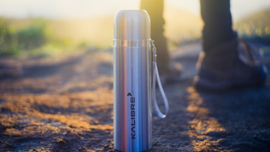A stainless steel thermos with the brand name 'KALIBRE' is positioned on a dirt path, with a strap attached to its side. In the blurred background, there are hints of sunlight and a pair of rugged boots, suggesting an outdoor setting with warm, natural lighting.