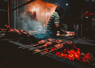 Chef carefully tending to meats on a glowing charcoal grill in a dimly lit kitchen.