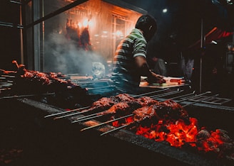 A person is grilling skewered meats over a barbecue with glowing coals. The scene is set in a busy and dimly lit area with smoke rising from the grill. Ambient lighting casts a warm glow, highlighting the cooking process.