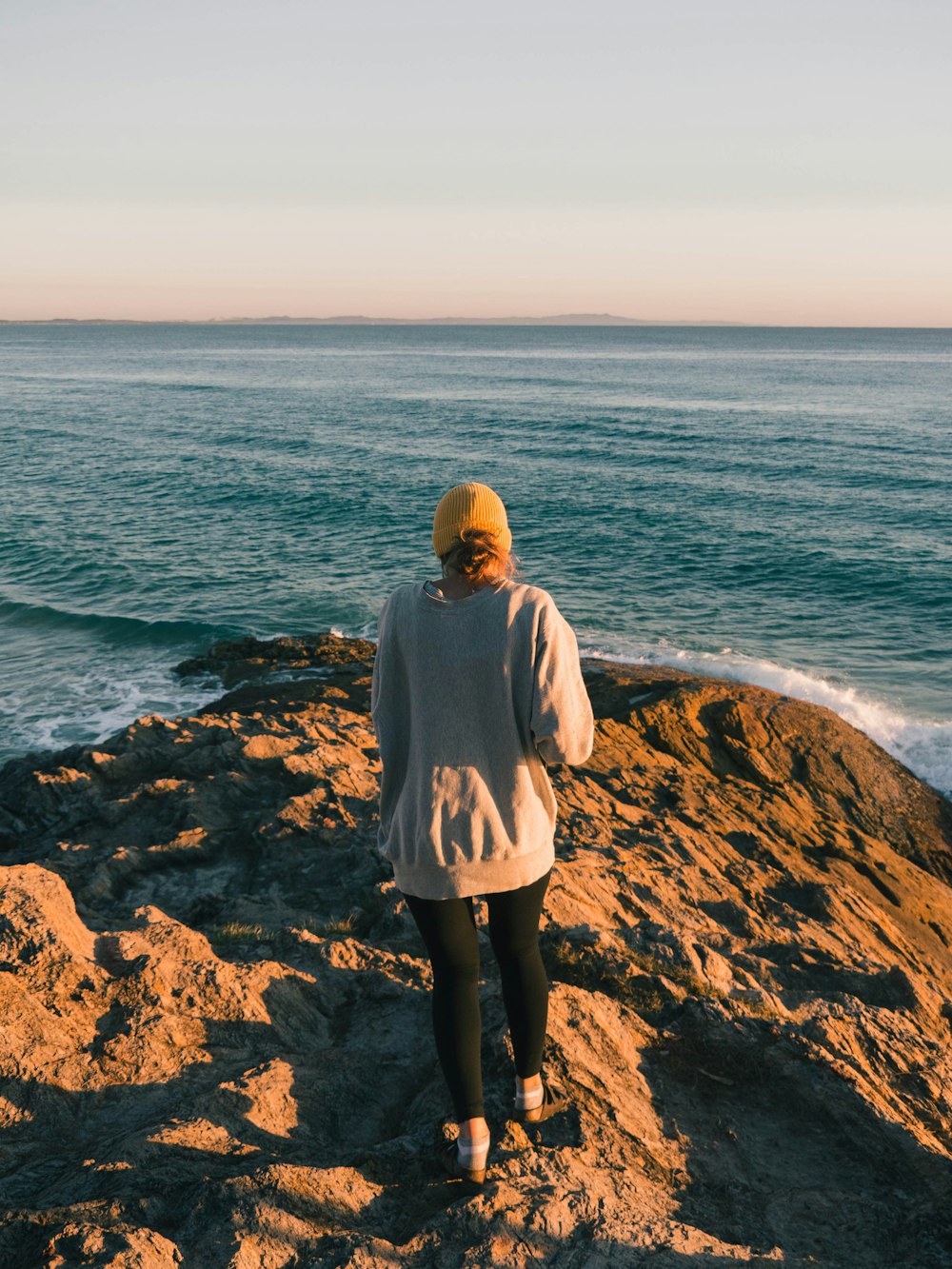 woman in gray hoodie standing on brown rock near body of water during daytime