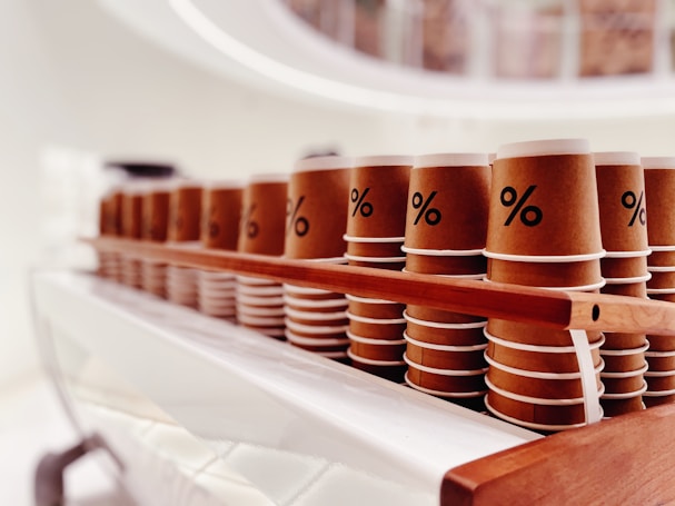A neatly arranged row of brown and white paper cups sits on a wooden tray, each with a prominent percentage symbol on the front. The background is softly blurred, suggesting a modern and minimalistic setting.