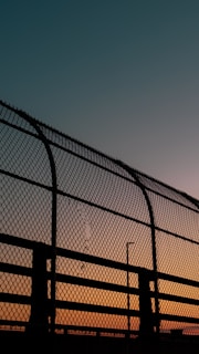 A chain-link fence silhouetted against a colorful sunset sky. The sky transitions from a deep turquoise at the top to a warm orange near the horizon. The fence, dark and prominent, features a damaged area where some links are broken.