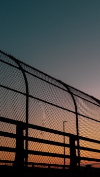 A chain-link fence silhouetted against a colorful sunset sky. The sky transitions from a deep turquoise at the top to a warm orange near the horizon. The fence, dark and prominent, features a damaged area where some links are broken.