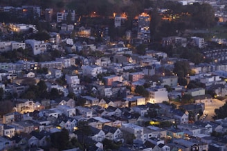 A densely packed urban neighborhood taken from an elevated perspective during the evening. The area features a mix of small to medium-sized residential buildings with varied architectural styles. Many homes have sloped roofs and are surrounded by trees. Warm lights emanate from the windows, contrasting with the dimming natural light. The landscape rises into a hill in the background, where more buildings are nestled among trees.