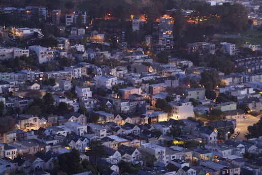 A densely packed urban neighborhood taken from an elevated perspective during the evening. The area features a mix of small to medium-sized residential buildings with varied architectural styles. Many homes have sloped roofs and are surrounded by trees. Warm lights emanate from the windows, contrasting with the dimming natural light. The landscape rises into a hill in the background, where more buildings are nestled among trees.