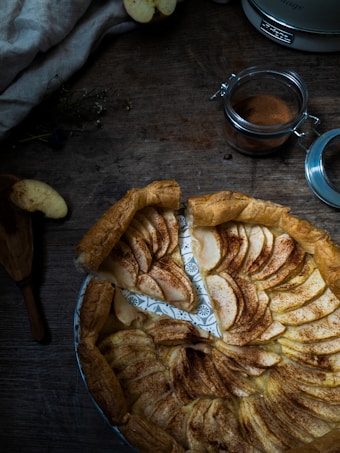 A wooden table with a freshly baked apple tart featuring cinnamon sprinkled apple slices arranged in a circular pattern on a rustic plate. A jar of cinnamon and a sliced apple are nearby, adding a touch of a cozy kitchen ambiance.