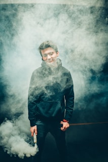 A person wearing a dark hoodie is surrounded by dense clouds of smoke, holding a canister labeled 'COLD SMOKE' in one hand. The setting appears to be dimly lit, enhancing the dramatic effect of the smoke.