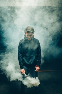 A person stands with a relaxed posture, holding a smoke stick that emits thick white smoke, creating an atmosphere limited by visibility. The individual is dressed in casual attire, including a dark hoodie, and appears to be in a dimly lit environment.