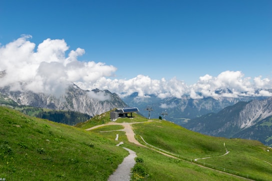 A serene mountain landscape featuring lush green hills, a small building with solar panels, and a pathway leading through the grass. Majestic mountains and a clear blue sky with scattered fluffy clouds create a breathtaking backdrop.