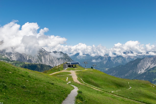 A serene mountain landscape featuring lush green hills, a small building with solar panels, and a pathway leading through the grass. Majestic mountains and a clear blue sky with scattered fluffy clouds create a breathtaking backdrop.