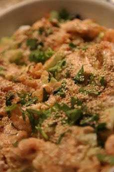A close-up view of a dish with shrimp or prawns covered in what appears to be a creamy sauce, garnished with sesame seeds and green herbs like cilantro or parsley. The focus is on the texture and details of the ingredients.