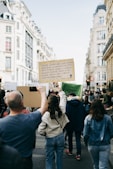 A group of people walking down a city street holding protest signs. One prominent sign quotes an environmental message. The street is lined with tall, classic European-style buildings. The atmosphere is dynamic with people actively participating in the protest.