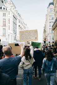 A group of people walking down a city street holding protest signs. One prominent sign quotes an environmental message. The street is lined with tall, classic European-style buildings. The atmosphere is dynamic with people actively participating in the protest.