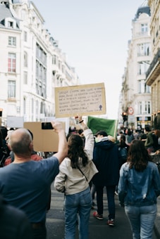 A group of people walking down a city street holding protest signs. One prominent sign quotes an environmental message. The street is lined with tall, classic European-style buildings. The atmosphere is dynamic with people actively participating in the protest.