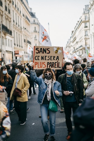 A group of people is gathered in a street, participating in a protest or demonstration. One person in the foreground is holding a sign with French text and a bee illustration. Many participants are wearing face masks and appear to be focused on the event. The surrounding architecture suggests an urban setting.