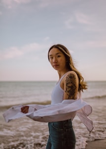 A laid-back model wearing a cream Bella Canvas tee with minimal wave line art, standing by the ocean shore at sunset.