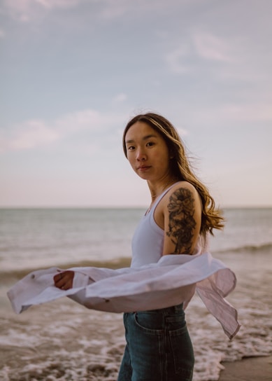 A laid-back model wearing a cream Bella Canvas tee with minimal wave line art, standing by the ocean shore at sunset.